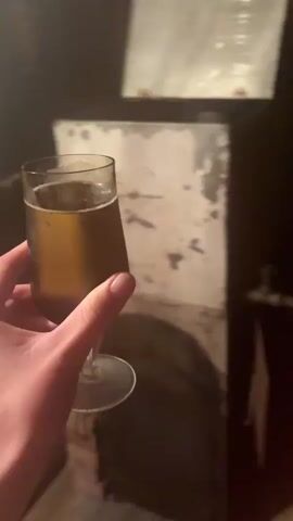 The video shows a person's hand holding a glass of amber-colored liquid, likely beer, with a stemmed glass. The background appears to be a wooden sauna or a similar environment, with wooden walls and a slatted wooden floor. A red plastic tub filled with green leafy material, possibly venik (a bath broom made of branches), is visible to the right of the glass. The lighting is dim, casting shadows on the wall. The camera movement is slightly shaky, suggesting it is being held by hand.