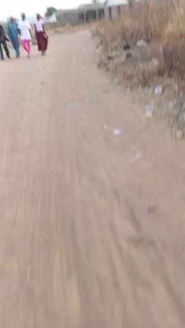 The video appears to be taken from a low vantage point, looking up a dirt path. Four people are walking away from the camera in the distance. The path is dusty, with some visible debris and tire tracks. The person recording seems to be moving forward, likely walking on the path as well. The camera shakes and points downward during the recording, blurring the view of the people in the distance. There are no discernable interactions or actions beyond walking. The environment seems rural or suburban, with sparse vegetation and a building visible in the background.