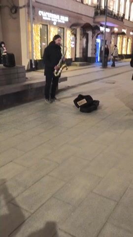 The video depicts a street scene at night. A man with long hair is standing and playing a saxophone on a city sidewalk. A small black amplifier is set on a ledge behind him, and an open black saxophone case with white lettering is on the ground in front of him. Pedestrians walk by, with one smiling directly at the camera. The buildings in the background have decorative lighting.