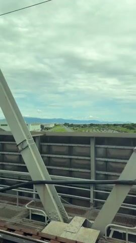 The video shows a view from a moving train. The train is traveling alongside a grey, paneled wall and a gravel track. Beyond the wall, there are green fields, a road, and some buildings. In the distance, there are mountains and a cloudy sky. The video passes by several structures, including industrial buildings and residential areas. A utility pole with wires and equipment is also visible. The overall impression is one of a rural or semi-rural landscape observed from a train.
