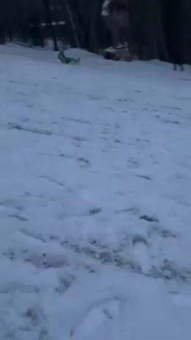 The video shows a snowy hillside. In the distance, a person on a sled is descending the hill. Two other people are standing further up the hill. Bare trees are visible in the background. The snow is marked with sled tracks and footprints. The sky appears overcast.