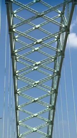 A view of a bridge from the perspective of a vehicle driving across it. The bridge has a distinctive green lattice structure supporting cables. The sky is blue with a few white clouds. Two cars are visible ahead on the bridge.