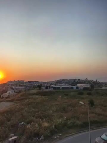 This is a video capturing a sunset over a coastal town. The sun, a bright orb of orange and yellow, is low on the horizon, casting a warm glow across the sky. The sky transitions from a soft orange near the sun to a pale blue at the top of the frame.

In the foreground, a grassy, unkempt hill overlooks a town. The town is characterized by a collection of buildings, many with white or light-colored walls and red-tiled roofs. Some of the buildings appear to be residential, while others look like apartment complexes or commercial structures. A road curves along the bottom of the frame, with a dark-colored car visible on it. The landscape is dry and sparsely vegetated, with tall grasses and small wildflowers. A lone lamppost stands on the hill, casting a faint shadow. The overall scene suggests a warm, possibly summer evening in Kuşadası, Turkey, given the coastal setting and architectural style.

The activity captured is the natural event of the sun setting, with the camera providing a static viewpoint of the panoramic scene. There are no people visible in the media. The notable details include the dramatic colors of the sunset, the silhouette of the town against the vibrant sky, and the arid vegetation of the hillside. The time of day is clearly sunset, and the weather appears clear and calm. The location cues strongly suggest Kuşadası, Turkey, a popular coastal resort town. The mood conveyed is one of peacefulness and natural beauty. There is no visible text within the media.