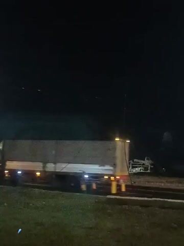 The video shows a nighttime scene from a moving vehicle. The primary focus is on a highway with several large trucks. One truck, a semi-trailer with a white tarp covering its cargo, is clearly visible in the foreground, moving from left to right. Its headlights and taillights are illuminated. In the background, a complex metal structure, possibly related to construction or infrastructure, is dimly lit. Other trucks and vehicles are present on the highway, some blurred due to motion. The environment appears to be outdoors, with a dark sky, suggesting it is late at night. The overall impression is of traffic on a highway at night, with a possible accident or unusual scene involving the metal structure in the distance.