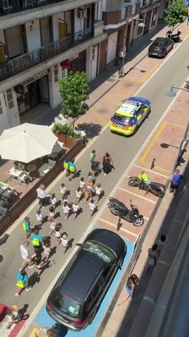A group of children, many wearing brightly colored shirts, are running down a street. They are accompanied by several adults. Some of the adults appear to be supervising the children, while others are walking alongside them. There are several parked cars on the street, and a police car is also present, with its lights flashing. The street appears to be in a town or city, with buildings lining both sides. Some of the buildings have shops on the ground floor. There are also umbrellas set up outside a cafe. The weather appears to be sunny. The children seem to be participating in some kind of organized activity, possibly a parade or a race. One adult is pushing a stroller. Another adult is holding a large umbrella over a group of children. The police car is stationary.
