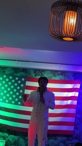 A man dressed as Elvis Presley stands in front of an American flag and sings into a microphone. He wears a white jumpsuit with silver buttons and embellishments. The room is illuminated with red, green, and blue stage lighting. There is a wicker chandelier hanging from the ceiling. The flag hangs against a backdrop of palm leaf wallpaper.