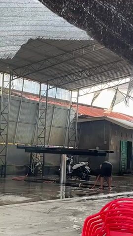 This media file shows an outdoor car wash facility in Tambonbaroh, Indonesia. The scene is set under a large, open-air metal roof structure covered with a mesh tarp, providing shade. The ground is wet and reflective, indicating recent washing activities.

In the foreground, bright red plastic chairs are stacked on the concrete floor. A person, wearing a dark t-shirt and shorts, is actively sweeping the wet ground with a broom. To the left, a white motorcycle is parked. Further back, under the roofed area, several black SUVs are visible. One SUV has its trunk open, revealing the interior. Another SUV is parked next to it, and a third is partially visible. The buildings in the background appear to be simple structures with corrugated metal siding and red-tiled roofs.

The lighting suggests it is daytime, possibly late afternoon or early morning, with the sun casting shadows and creating bright highlights on the wet surfaces. The overall atmosphere appears to be a typical day at a car wash, with the focus on cleaning vehicles and maintaining the facility. No specific weather conditions are apparent beyond the presence of water on the ground, which is expected for a car wash. The visible text on the license plates of the cars includes "BL 1239 KK" and "BL 1828 Y".
