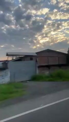 The video shows a view from inside a moving vehicle. The outside scenery consists of a building, vegetation, and a cloudy sky with the sun peeking through. The building has a red brick exterior. The vegetation is primarily green grass and trees. The sky is blue with scattered white and gray clouds. At one point, a yellow directional arrow road sign is visible.