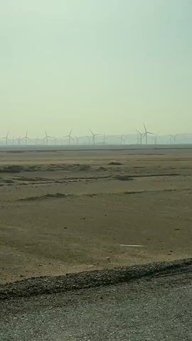 The video shows a wide, flat, arid landscape in hazy conditions. A distant mountain range is barely visible on the horizon. In the foreground, the terrain is dry and cracked, with a few darker patches that suggest dampness or different soil composition. The main feature of the scene is a large wind farm in the middle distance, consisting of numerous wind turbines. The turbines are spread across the landscape, some more clearly visible than others due to the haze. The camera seems to be moving, suggesting that the video was taken from a moving vehicle. The overall impression is that the video depicts a desolate but technologically advanced environment.