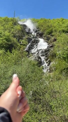 The video displays a waterfall cascading down a rocky hillside covered in lush green vegetation. The water appears white and frothy as it falls, creating a mist at the top of the falls. The sky is a bright blue with a few white clouds. A tall metal tower is visible on the top of the hill behind the waterfall. A hand briefly appears in the lower left corner of the screen.