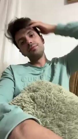 A man is sitting on a chair with a textured pillow in his lap. He is wearing a light teal sweater and has dark hair. He opens a tube of hand cream, squeezes some into his hand, and pauses.