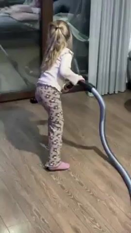 A young girl, seen from the back, is vacuuming a wooden floor in a room. She has blonde hair tied in a ponytail. She is wearing a light-colored, long-sleeved shirt and printed leggings, along with pink slippers. She is holding a gray vacuum hose and moving it back and forth in front of a large glass door. The glass door reflects the room's interior, including a vacuum cleaner resting on the floor. The floor is made of light-colored wood planks. White curtains are partially visible in the background, to the right of the girl. The overall lighting of the room appears bright and natural.