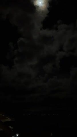 The media file captures a nighttime scene with the moon partially obscured by dark, textured clouds. The moon itself is a bright, luminous orb, casting a soft glow through the atmospheric haze. Below the sky, a vast expanse of darkness suggests a landscape or body of water. A faint line of distant lights stretches horizontally across the lower portion of the frame, hinting at a settlement or a coastline.

In the bottom left corner, the edge of what appears to be a structure or object is visible, with a bright light reflecting on its surface. This could be part of a boat, a building, or an elevated platform. The overall impression is one of a tranquil, yet somewhat mysterious, night.

The time of day is clearly night, indicated by the darkness and the presence of the moon. The weather appears to be partly cloudy, with the clouds playing a significant role in framing the moon. The location context of Ko Tao Subdistrict, Thailand, suggests a tropical setting, though this is not explicitly visible in the image itself, other than perhaps the faint lights suggesting coastal activity or island life. The emotions evoked are likely to be a sense of wonder, peace, and perhaps a touch of awe at the celestial display. No text is visible in the media file.