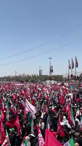The video shows a large gathering of people in an open outdoor area under a clear blue sky. The crowd is densely packed, and many individuals are holding flags of various colors, predominantly red, green, and white, which appear to be the colors of the Iranian flag, along with other red flags bearing Arabic script. In the background, a distinctive tower-like structure with a geometric design is visible, along with buildings and trees, suggesting an urban environment. The people in the crowd are mostly wearing dark clothing. The overall scene depicts a large public assembly or demonstration.