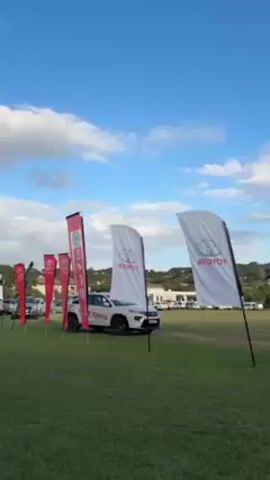 The video showcases an outdoor event hosted by Toyota. The scene opens with flags displaying the Toyota logo placed in a grassy field, adjacent to Toyota vehicles. A man is seen walking in the background. Subsequently, the camera pans to a yellow Toyota vehicle with "Kowie Toyota" branding, parked on the field. The video ends with a wide shot of the event, showing a large tent set up with people gathered underneath, sitting at tables. The environment is sunny with a partly cloudy, blue sky and a green, hilly landscape in the distance.