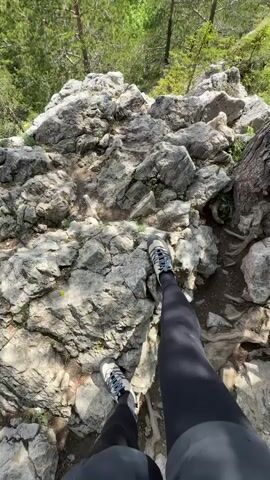 The video opens with a first-person perspective looking down at a person's feet in hiking shoes, standing on a rugged, rocky mountain trail. The path is uneven, composed of large grey and brown stones, with some roots and patches of dirt visible. Lush green trees flank the trail.

The camera then pans up, revealing a breathtaking panoramic view of a vast mountain landscape. Below the rocky viewpoint, dense, vibrant green forests stretch across rolling hills and valleys. In the distance, majestic mountain ranges with hints of snow on their peaks rise under a partly cloudy sky. The clouds are a mix of white and grey, suggesting a dynamic weather pattern but with good visibility.

Pine trees, some with exposed roots, frame the immediate foreground, adding to the natural, untamed feel of the environment. The sunlight highlights the different shades of green in the foliage and the texture of the rocks. The setting is clearly a high vantage point, offering an expansive view over what appears to be the Tatra Mountains in Zakopane, Poland, known for their dramatic peaks and extensive forests. The overall impression is one of serene natural beauty and the exhilaration of being in the mountains.