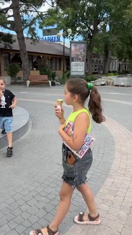 Two young girls are walking on a paved path in front of a fountain and a building. Both girls are holding and eating ice cream cones. The girl in the foreground is wearing a neon green top and denim shorts. She is holding a book in her left hand. The girl in the background is wearing a black t-shirt and denim shorts. She is also holding an ice cream cone. The fountain is ornate and made of stone. The building behind them appears to be a restaurant or shop with outdoor seating. There are trees and other buildings visible in the background. The weather appears to be overcast.