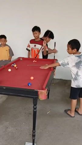 Five children are gathered around a red pool table. One child in a white and red shirt is actively playing, holding a cue stick and positioned to strike a ball. Another child in a black shirt stands behind him, also holding a cue. Two other children are observing to the left, one in a yellow and grey shirt and the other in a black t-shirt and shorts. A fifth child in a black and white tie-dye shirt is on the right, holding a cue stick. The pool table has several colored balls on it. The scene appears to be indoors, with a light-colored wall and a concrete floor.
