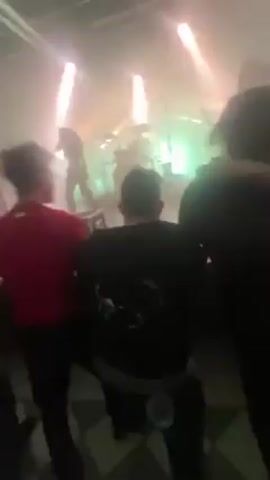 The video shows a band performing on a stage in a dimly lit venue, likely a concert. The stage is illuminated by several bright lights, some of which are white and green. There is a noticeable amount of smoke or haze in the air, contributing to the overall low visibility. Several people are in the foreground, their figures mostly obscured, suggesting the perspective of someone in the audience. The camera moves, focusing on the stage and then briefly shifting towards the ceiling, which reveals lighting and sound equipment. The overall impression is that the viewer is experiencing the concert atmosphere from within the crowd.