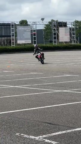 A person on a red and white motorcycle is practicing maneuvers in a large, empty parking lot. The lot is paved and marked with white lines, suggesting it may be used for training or events. Orange cones are visible on the ground, likely part of the training course. The rider is wearing a full motorcycle suit, including a helmet and gloves, indicating safety precautions are being taken. In the background, there are buildings, trees, and a few parked vehicles, including a white van. The sky is overcast. The motorcycle appears to be a smaller, perhaps off-road or pit bike style, with knobby tires. The rider is leaning into a turn, demonstrating control and skill on the bike.
