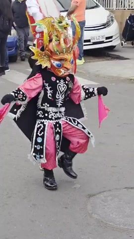 The video shows a person in a colorful costume dancing in the street. The costume includes a large, ornate mask that covers the wearer's face, resembling a dragon or mythical creature with orange, blue, and gold accents. The costume also features a black cape with white trim and dangling fringes, a black vest adorned with white swirling patterns and blue circular embellishments, and bright pink pants and sleeves. The person is wearing black, shiny boots and black gloves. They are holding two pink fabric streamers, one in each hand, which they appear to be using in their dance. The dance involves jumping and arm movements. In the background, there are several people standing on the sidewalk, some of whom are watching the dancer. A white car is parked on the street behind the dancer. The scene appears to be outdoors, possibly during a parade or festival, on a paved street with yellow lines marking the edge of the roadway. The lighting suggests it is daytime.