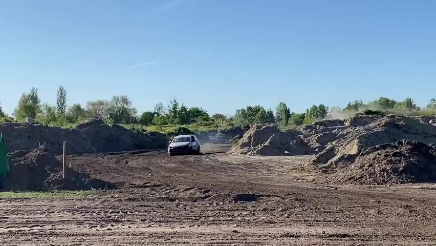 The video shows a dirt track racing event. Three cars, one silver, one white, and one red, are racing around the track, kicking up dust and dirt. The track is surrounded by dirt piles and some greenery. The sky is clear and blue. A green flag is visible on the left side of the frame.