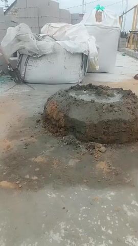 The video shows an outdoor construction or work site. In the foreground, a circular mound of wet concrete or cement is visible. Behind the concrete, there are large white bags, possibly containing building materials like cement or sand, covered in plastic sheeting. To the right of the concrete, a man wearing a cap and a t-shirt is sitting on a low wall, holding a small object, possibly a tool or a drink. A black bucket is also on the ground near him. In the background, stacks of concrete blocks are visible, and overhead power lines suggest an industrial or semi-urban setting. The ground appears to be paved, with some dirt and scattered debris. The overall impression is a work in progress.