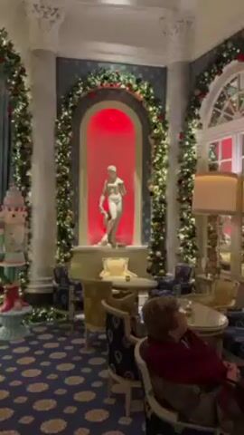 The video shows the interior of a room decorated for Christmas. The room features a blue carpet with a floral pattern, ornate white columns and archways adorned with garland and string lights. There are several Christmas trees, some decorated with ornaments and lights. A statue stands in a niche illuminated with red light. Seating areas include chairs and small tables. A woman is sitting in one of the chairs. The camera pans slowly across the room, showcasing the decorations and furnishings.