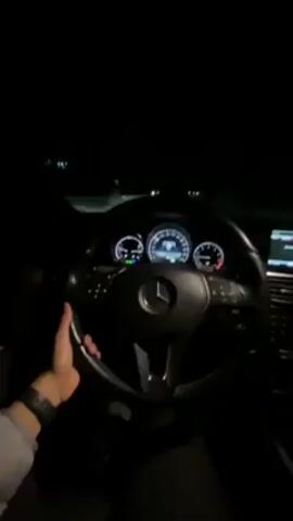 Here's a description of the video:

A first-person perspective shot from inside a Mercedes-Benz at night. The dashboard and steering wheel are prominently visible, illuminated by the car's interior lights. A person's hand wearing a smartwatch is visible on the steering wheel. The car appears to be moving on a dark road. The dashboard displays the car's speed and other information.