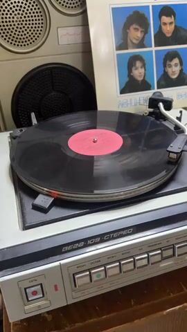A turntable plays a black vinyl record with a red label. The player is gray and marked "VEGA-109 STEREO". Next to it, a record sleeve displays a band with four members. A speaker with a grill is visible in the background. The camera angle remains static and provides a close view.