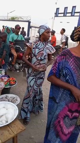 The video shows an outdoor marketplace with vendors and customers. A woman in a blue patterned dress and headscarf stands in the foreground. There are several trays of food items displayed on tables, including what appears to be yams, tomatoes, onions, and fish. People are walking around, some are on motorcycles. A man in a yellow shirt with a tiger stripe pattern is seen interacting with a young boy. The environment appears to be a busy public space with buildings visible in the background.