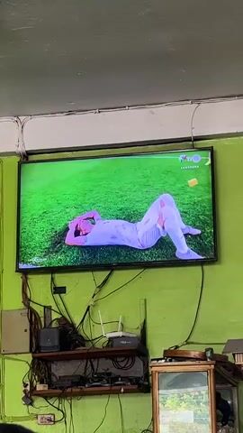 A large flat-screen television mounted on a green wall displays a soccer match. The screen shows a player lying on the green grass, wearing a white and pink uniform. Another player in a yellow and blue uniform approaches and kneels beside him. Other players and spectators are visible in the background on the screen. Below the television, a shelf is cluttered with electronic devices, wires, and a router. To the right, a wooden cabinet with a glass door is visible. The lighting is dim.