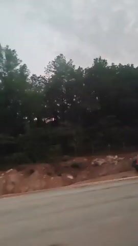 The video shows a cloudy sky with a hint of pink near the horizon, suggesting either sunrise or sunset. A dark silhouette of trees and hills is visible against the sky. In the foreground, there is a road, possibly asphalt, with a white line on the right side. The road appears to be wet, reflecting the sky. A thin, vertical pole or shadow is visible on the road. The overall impression is that the video was captured from a moving vehicle, likely a car, as it travels along the road.