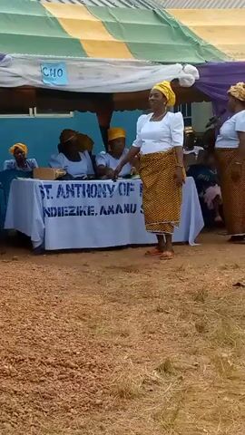 The video shows two African women singing and dancing outdoors in front of a table covered with a white tablecloth that reads "ST. ANTHONY OF NDIEZIKE AMAMU." The women are wearing white tops, yellow head wraps, and yellow-and-brown patterned skirts. One woman is holding a microphone. Behind the table, other women are seated, also wearing yellow head wraps. A tent with green, yellow, and purple fabric is visible overhead. The ground is dry with scattered straw.