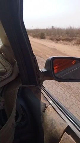 The video shows a view from inside a car driving on a dusty road. The car's interior is worn and rusted. The windshield is cracked and there is a backpack on the dashboard. Outside, the landscape is arid with sparse vegetation. The air is hazy with dust, reducing visibility. The car is moving along a paved road, with unpaved shoulders. Another vehicle can be seen in the distance through the dust. There is a sticker on the windshield referencing the Joint Tax Board.