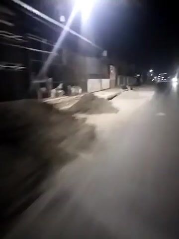 The video shows a view of a road at night, seemingly taken from a moving vehicle, potentially a bike or motorcycle. The road is asphalt and appears to be illuminated by the vehicle's headlights, as well as streetlights in the distance. There's construction material, possibly sand or dirt, piled up along the side of the road. Buildings are visible on the roadside, with some appearing to be closed businesses or residences. The overall atmosphere is dark and quiet, with limited traffic.