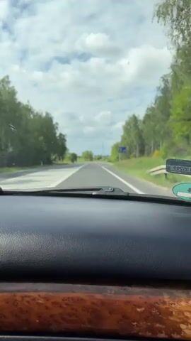 A view from inside a vehicle traveling on a rural road. The dashboard is black with a faux wood trim. The road has white lane markings and an arrow directing drivers to the left. The sky is cloudy and there are trees lining the road.