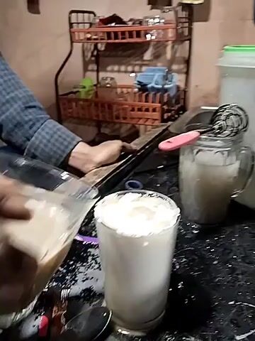 A man is pouring a liquid from a square glass into a clear glass full of white cream. The glass with cream is sitting on a black countertop. Another glass full of a cloudy liquid, and a white plastic container, are sitting nearby. A metal whisk with a pink handle rests in the second glass. Behind the man is a brown metal wire rack with assorted items on it. The wall behind the rack is cream colored and has some small items stuck to it. The man is wearing a blue and white plaid shirt. The light source is dim and the video quality is grainy.