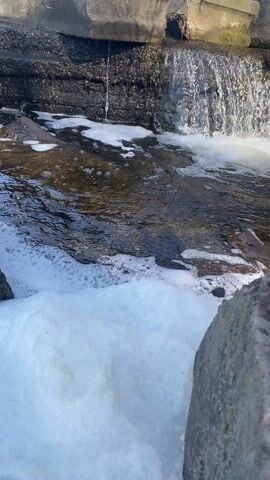 The video shows a small waterfall cascading over dark rocks into a stream. The water churns and creates white foam in the pool at the bottom of the falls. A large grey rock is visible in the foreground on the right. The overall scene appears to be a close-up of a natural or landscaped water feature.