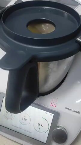 The video shows a close-up shot of the top of a kitchen appliance, likely a food processor or blender. A dark grey lid with a circular opening is prominently featured. Through the opening, a light yellow, creamy substance is visible, swirling and churning. The appliance itself appears to be white with a touchscreen interface displaying numerical values and a circular graphic. The overall environment suggests a domestic kitchen setting. The video captures the action of the food processor in operation, processing the yellow substance.