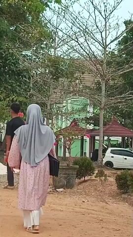 Two women wearing hijabs walk away from the camera on a dirt path. The woman in the foreground is wearing a black long-sleeved shirt and a grey hijab. She is holding a phone in her right hand. The woman in the background is wearing a pink and white patterned dress and a grey hijab. She is carrying a dark-colored bag in her left hand. To the left of the women, a young man wearing a black t-shirt and dark pants walks away from the camera. He is holding a red object in his right hand. In the background, there are trees and a light green building with a dark roof. The ground is covered in dirt and some scattered white material. The sky is overcast.