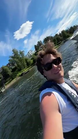 The video begins with a close-up of a young man wearing sunglasses and a white t-shirt, sitting in the front of a rigid inflatable boat. He is smiling and holding a phone, likely recording a selfie. Behind him, another person, wearing a blue baseball cap and a white t-shirt, is steering the boat. They are both wearing life vests. The boat is moving through choppy water, creating a white wake. The camera pans to show the surroundings: a body of water with other boats visible in the distance, a shoreline with trees and houses, and a clear blue sky with some clouds. The second half of the video shows more of the scenery, including a closer view of modern houses built on a hillside overlooking the water. The sun is shining brightly.