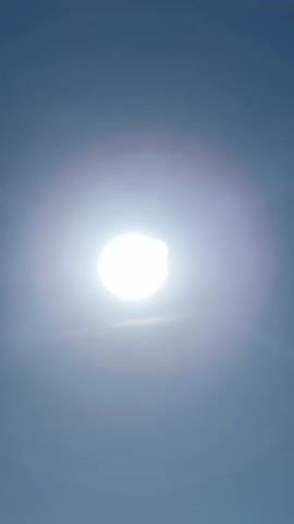The video shows a bright sun against a clear blue sky. The sun is in the center of the frame, and its light creates a lens flare effect. There are some small dust or water particles visible in the air.