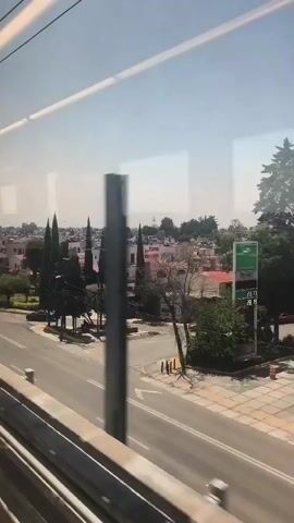 The camera, located inside a moving train, captures an exterior view through the window. The primary focus is on a street scene outside the train, featuring a typical residential area.

The street is visible with light vehicle traffic, including cars and a yellow van. Sidewalks line the street, bordered by landscaped greenery. Beyond the street, the view expands to reveal a cluster of white houses with red roofs, typical of residential neighborhoods. Trees of varying sizes dot the landscape.

The sky is clear and bright, suggesting a sunny day. The metallic supports inside the train are visible in the foreground. The camera's movement creates a dynamic perspective as the train travels through the area.
