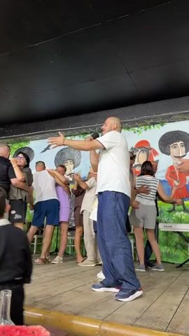 A man with a bald head is holding a microphone and singing in front of a group of people who are dancing. He is wearing a white t-shirt and blue jeans. The dancers are of various ages and genders, and they are all dressed in casual clothing. The background of the scene is a wall with a mural depicting people in traditional clothing. The floor is made of wood. In the foreground, there is a table with a glass and some watermelon slices. The lighting appears to be artificial, possibly from stage lights.