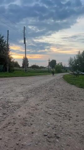 A young boy rides a bicycle down a dirt road. The sun is setting in the background, casting a warm glow over the scene. The road is unpaved and appears to be in a rural area, with trees and houses visible on either side. The sky is filled with dramatic clouds. Another person, possibly an adult, is visible further down the road to the right, walking away from the camera. The boy is wearing a yellow t-shirt and dark pants. The bicycle is red and has visible spokes and pedals. The overall mood of the video is peaceful and serene.