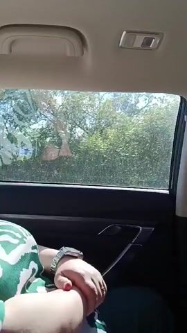 The video shows a view from inside a car traveling through a rural area in Belarus. The scene viewed through the car window is dominated by lush green vegetation, including trees and tall grasses, with a body of water, likely a lake or river, visible in the distance. The sky is bright with scattered clouds, suggesting a sunny day. The perspective is from the back seat, with a person's arm and shoulder visible in the foreground. They are wearing a green and white patterned top and a wristwatch. The interior of the car is also visible, including a door handle and the ceiling with lights and a grab handle. The overall mood is peaceful and scenic, indicative of a leisurely drive through the countryside. There is no visible text in the media file.