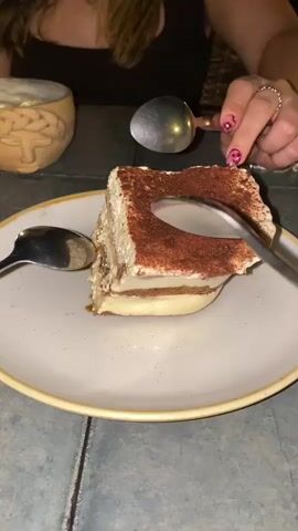 Here's a description of the video:
A person is cutting into a piece of tiramisu on a white plate with a yellow rim. They use two spoons to break off a piece, separating the cocoa-dusted top from the creamy filling and ladyfingers. The person wears a dark top and has a ring on their finger. One hand has pink-and-white nail polish while the other has red nail polish. The table has a gray-green color and a textured surface. Beside the plate is a container that resembles a brain. In the background, music is playing.