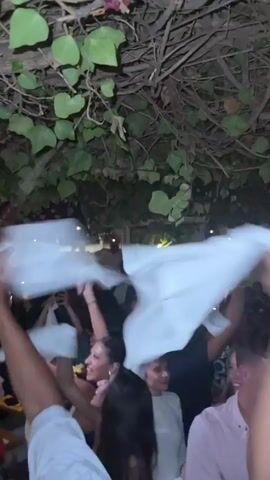 A group of people are at a party, possibly a celebration, with many holding white cloths or napkins and waving them in the air. The scene is outdoors or in an open-air venue, with a canopy of green leaves and vines overhead. Some people are dancing and appear to be enjoying themselves. There's a bar area visible in the background with bottles on display. The lighting suggests it is evening.