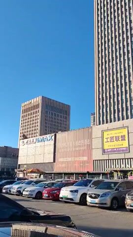 The video shows a street view with a tall building and a parking lot in front of it. Several cars are parked in the lot, and some people are walking around. The building appears to be a commercial building, with shops and offices. The sky is clear and blue. The video is shot from a low angle, looking up at the building. The lighting suggests it is daytime, likely sunny. The overall scene depicts an urban environment.